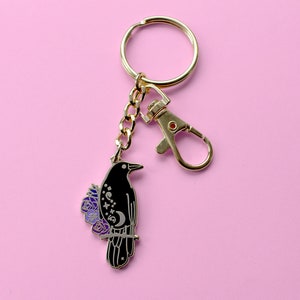 May include: Gold key chain with a black crow charm with purple flowers and a crescent moon design.