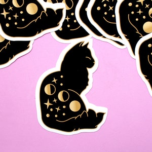 May include: A black cat silhouette sticker with gold moon and star details. The cat is curled up in a crescent moon shape.