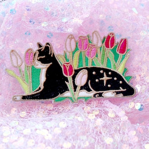 May include: Embroidered patch depicting a black cat with white markings, nestled amongst pink and red tulips with green leaves. The cat has a starburst design on its back. The patch is set against a pink, sparkly backdrop.
