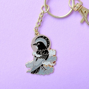 May include: A gold-toned metal keyring featuring a black and grey enamel raven perched amidst swirling white and light blue glitter clouds, set against a crescent moon background. The charm is attached to a circular ring and a lobster clasp.