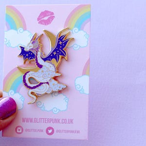May include: Purple and white enamel pin of a dragon curled around a crescent moon with stars. The dragon has a white body with purple accents and a gold outline. The background is a pink card with a rainbow and clouds.