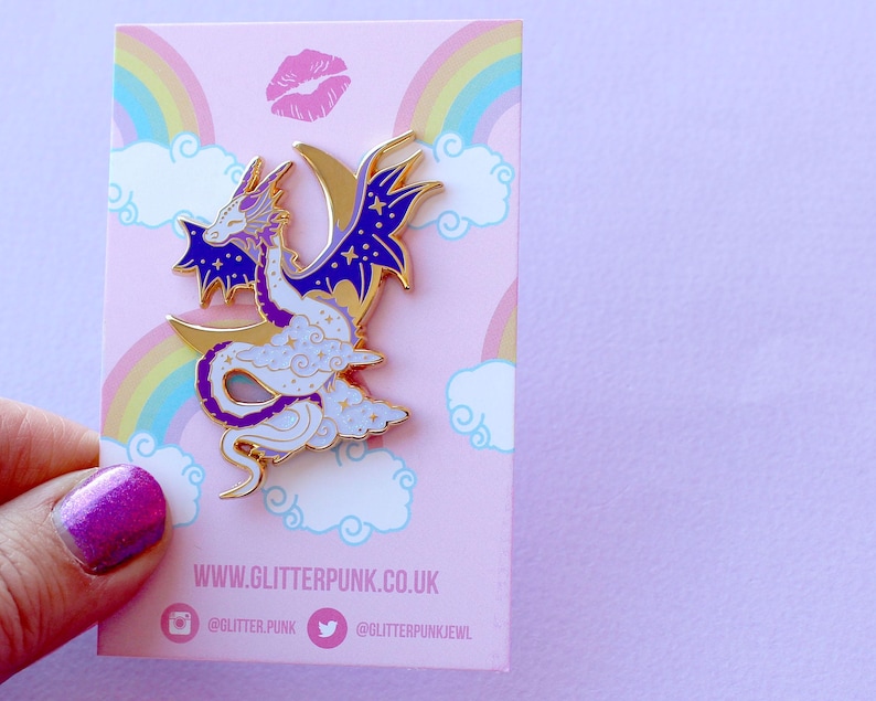 May include: Purple and white enamel pin of a dragon curled around a crescent moon with stars. The dragon has a white body with purple accents and a gold outline. The background is a pink card with a rainbow and clouds.