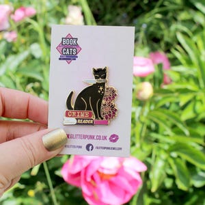 May include: A black cat enamel pin with a knife, the words "Crime Reader" on a book, and "Book Cats". The pin has gold accents and is on a white card. The website address w.glitterpunk.co.uk is visible.