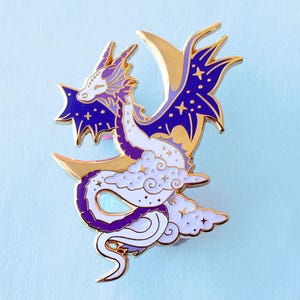 May include: A purple and white enamel pin of a dragon curled around a crescent moon. The dragon has a white body with purple accents and a gold crescent moon. The dragon's wings are spread out and have a purple and gold design.