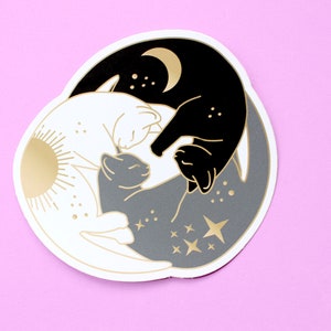 Pu&ograve; includere: Un adesivo fustellato con un design Yin e Yang con gatti. Il design include un gatto nero con una luna dorata, un gatto bianco con un sole dorato e un gatto grigio con stelle dorate. L'adesivo ha un bordo bianco ed &egrave; posto su uno sfondo rosa.