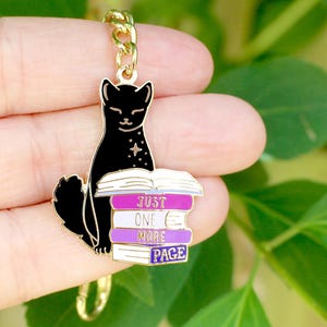 May include: A black cat charm with a gold chain. The cat is sitting on a stack of books with the text "JUST ONE MORE PAGE" in white and purple. The charm is likely a keychain or pendant.