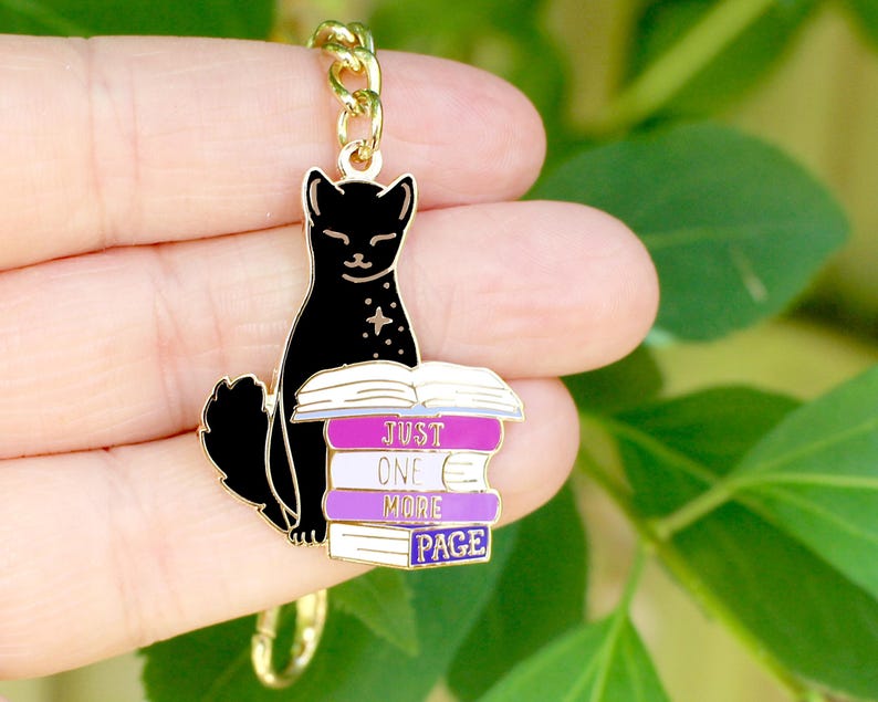 May include: A black cat charm with a gold chain. The cat is sitting on a stack of books with the text "JUST ONE MORE PAGE" in white and purple. The charm is likely a keychain or pendant.