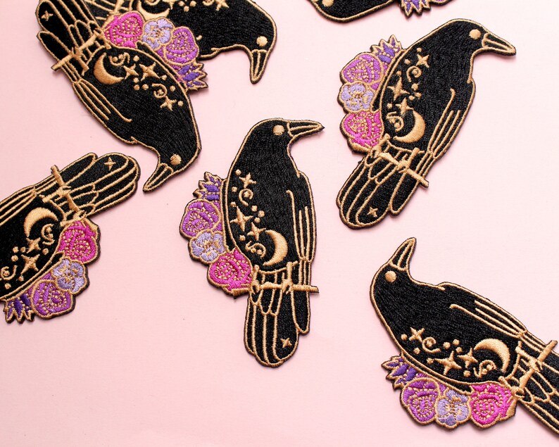 May include: Embroidered black raven patches with gold accents, featuring celestial designs and floral details. The patches showcase a crescent moon, stars, and flowers in shades of pink and purple. These patches are ideal for adding a unique touch to clothing or accessories.