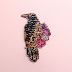 May include: Embroidered raven patch with black, gold, and silver thread. The raven has a floral design with pink, purple, and gold accents. The patch is likely for clothing or accessories.
