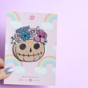 May include: A brown embroidered iron-on patch of a smiling pumpkin wearing a flower crown with pink, blue, and white flowers. The patch is on a pink background with rainbow and cloud designs. The text "www.glitterpunk.co.uk" is at the bottom of the image.