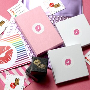 Pu&ograve; includere: Una collezione di scatole rosa e bianche con un adesivo con stampa di labbra rosa, insieme a biglietti da visita e una carta a righe arcobaleno. Le scatole sono disposte su una superficie rosa. I biglietti da visita riportano il testo "Glitter Punk".