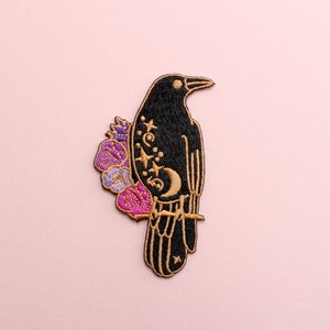 May include: Embroidered patch featuring a black raven perched on a branch, adorned with gold accents and a crescent moon. Purple and pink flowers embellish the raven's back. The patch is set against a soft pink background, ideal for jackets, bags, or other fabric items.