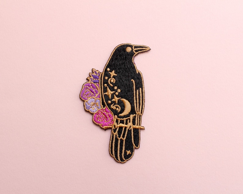 May include: Embroidered patch featuring a black raven perched on a branch, adorned with gold accents and a crescent moon. Purple and pink flowers embellish the raven's back. The patch is set against a soft pink background, ideal for jackets, bags, or other fabric items.
