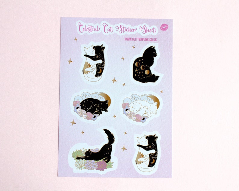 May include: A sheet of stickers featuring celestial cat designs. The stickers are on a light purple background with gold stars. The designs include a black cat with a crescent moon and stars, a white cat with a sun and stars, a black cat sleeping with flowers, a white cat sleeping with flowers, a black cat stretching with flowers, and a white cat hugging a black cat with a sun and moon.