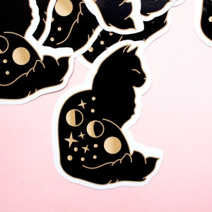 May include: Black and gold cat sticker with a crescent moon and stars design. The cat is sitting with its tail curled around its body.