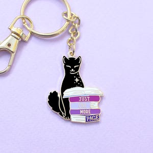 May include: A gold-colored keychain with a black cat design. The cat sits atop a stack of books with the text "JUST ONE MORE PAGE" in white. The keychain has a lobster clasp and a split ring.