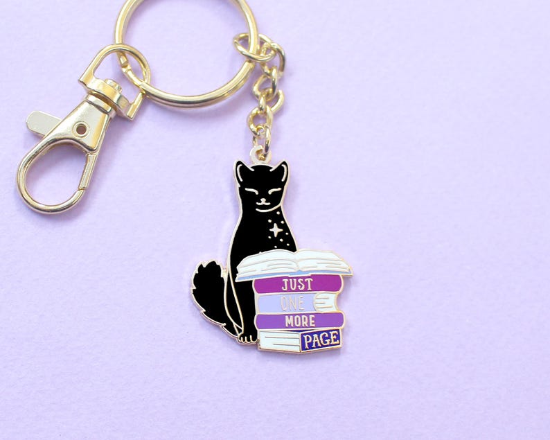 May include: A gold-colored keychain with a black cat design. The cat sits atop a stack of books with the text "JUST ONE MORE PAGE" in white. The keychain has a lobster clasp and a split ring.