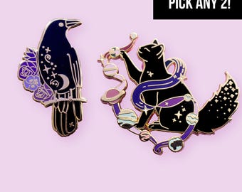 May include: Two enamel pins featuring a black crow with purple flowers and celestial symbols, and a black cat surrounded by planets and stars. The image includes a black rectangular box with the white text PICK ANY 2! in the upper right corner.