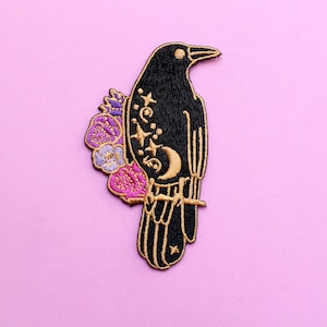 May include: Embroidered patch featuring a black raven perched on a branch, adorned with gold accents, stars, and a crescent moon. The raven is embellished with purple and pink flowers, set against a light pink background. This patch is suitable for clothing or accessories.
