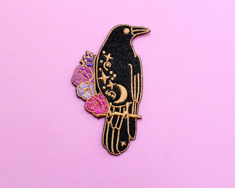 May include: Embroidered patch featuring a black raven perched on a branch, adorned with gold accents, stars, and a crescent moon. The raven is embellished with purple and pink flowers, set against a light pink background. This patch is suitable for clothing or accessories.