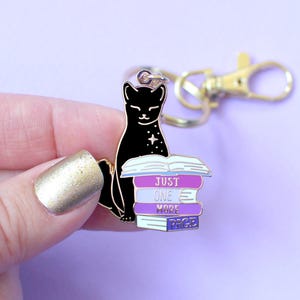 May include: A black cat keychain with a gold clasp. The cat is sitting on a stack of books with the words "JUST ONE MORE PAGE" in white. The books are purple, blue, and white. The background is a light purple.