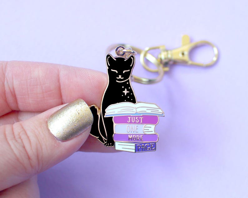 May include: A black cat keychain with a gold clasp. The cat is sitting on a stack of books with the words "JUST ONE MORE PAGE" in white. The books are purple, blue, and white. The background is a light purple.