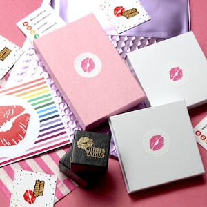 May include: Pink, white, and black boxes with a lip design and the text "Glitter Punk" on them. The boxes are on a pink surface with rainbow striped paper and a white and pink polka dot paper.
