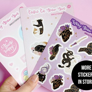 May include: Three sticker sheets with various designs. The first sheet features a pink background with a white sticker that says "Think happy". The second sheet features a purple background with a celestial cat design. The third sheet features a purple background with a witchy design, including a black cat, a snake, a cauldron, and a heart.