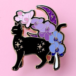 Peut inclure: Épingle en émail représentant un chat noir avec des bordures dorées, orné de fleurs violettes et bleues, d'un croissant de lune et d'étoiles. Le chat a des marques en forme d'étoiles. Sur fond rose.