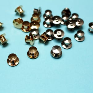 May include: A close-up of a group of silver and gold safety pins. The pins are scattered on a light blue surface.