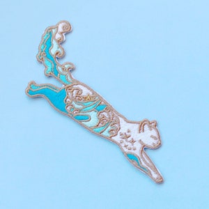 May include: A white cat with blue and gold wave patterns embroidered on its body, leaping with its tail curled up in a wave shape. The cat is on a light blue background.