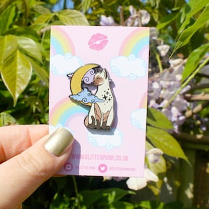 May include: An enamel pin featuring a cat on a crescent moon, with clouds and a rainbow on a pink background. The pin has the website www.glitterpunk.co.uk. The cat is white, brown, and black.