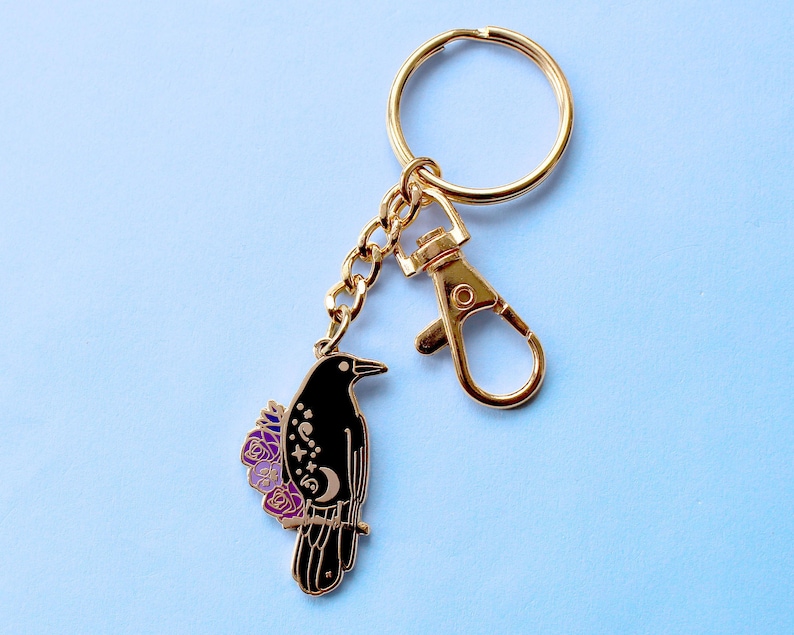 May include: Gold key chain with a black crow charm. The crow has purple flowers and celestial designs on its body.