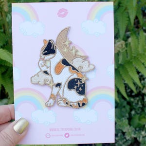May include: Embroidered patch of a calico cat with a moon and stars design. The patch is on a pink card with a rainbow and cloud pattern. The card also has the website address www.glitterpunk.co.uk.