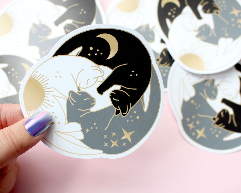 Pu&ograve; includere: Adesivi rotondi con un design yin-yang con gatti. Un lato &egrave; nero con una luna crescente, l'altro bianco con un sole. Un gatto grigio &egrave; al centro, con stelle e dettagli dorati. Gli adesivi sono tenuti da una mano.