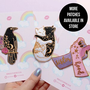 May include: Embroidered patches featuring a black raven with floral accents, two hugging cats, and a hand holding a ribbon with the words "sisterhood". The patches are on a pink background with rainbows and clouds.