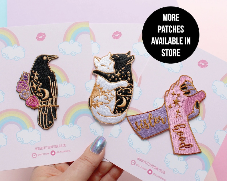 May include: Embroidered patches featuring a black raven with floral accents, two hugging cats, and a hand holding a ribbon with the words "sisterhood". The patches are on a pink background with rainbows and clouds.