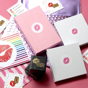 May include: Pink, white, and black boxes with a lip design and the text "Glitter Punk" on them. The boxes are on a pink surface with rainbow striped paper and a white card with a lip design and the text "Glitter Punk".