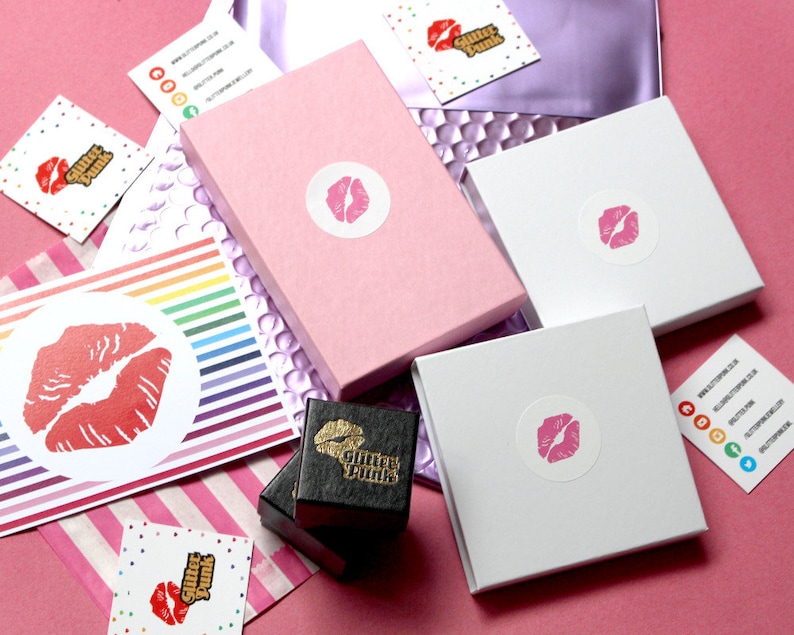 May include: Pink, white, and black boxes with a lip design and the text "Glitter Punk" on them. The boxes are on a pink surface with rainbow striped paper and a white card with a lip design and the text "Glitter Punk".