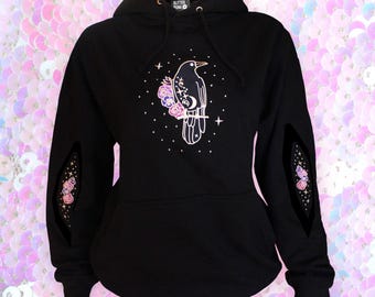 May include: Black hoodie with a crow and floral design. The graphic features a crow perched on a branch with flowers and stars. The sleeves have decorative cutouts with floral and star accents. The hood has the text "Glitter Punk".