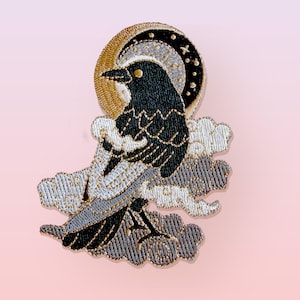 以下が含まれることがあります： 金と黒の三日月の前に配置された黒いカラスの刺繍ワッペン。鳥は、複雑なステッチのディテールが施された様式化された白と灰色の雲に囲まれており、層状の天体のデザインを作り出しています。
