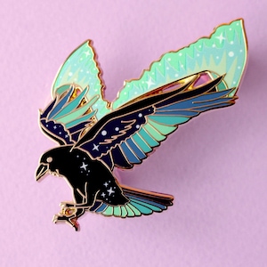 Könnte beinhalten: Detaillierte Emaille-Anstecknadel, die einen Raben mit ausgebreiteten Flügeln darstellt. Der Rabe ist schwarz mit einem Sternenmuster auf Flügeln und Körper. Die Flügel weisen Blau- und Türkistöne mit goldenen Konturen auf, vor einem hellvioletten Hintergrund.