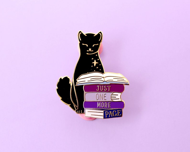 Op de afbeelding: Zwarte kat emaille pin met een stapel boeken waarop "Just One More Page" staat.