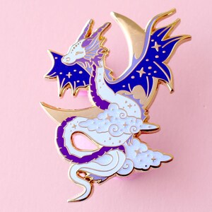 May include: Gold-tone enamel pin featuring a white dragon with purple accents, curled around a crescent moon with stars and clouds.
