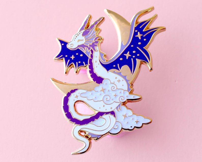 May include: Gold-tone enamel pin featuring a white dragon with purple accents, curled around a crescent moon with stars and clouds.