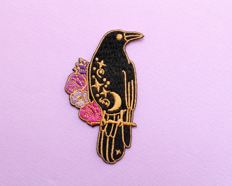 May include: Embroidered black raven patch with gold outline, perched on a branch. The raven features a crescent moon, stars, and floral accents in purple and pink. The patch is set against a light purple background.