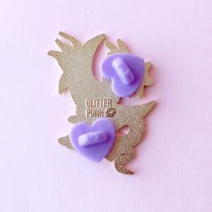 May include: Gold-colored enamel pin featuring a cartoon dragon with a purple heart on its chest. The pin has two purple butterfly backings and the text "Glitter Punk" on the back.