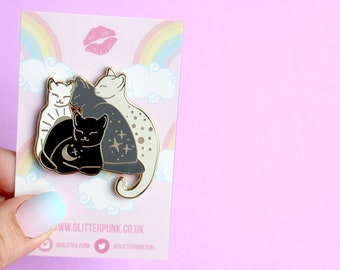 May include: Gold enamel pin featuring three stylized cats huddled together. The cats are black, white, and gray with celestial details. The pin is on a white card with a rainbow and clouds.
