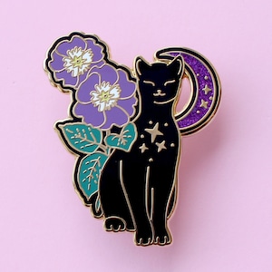 Peut inclure: Épingle en émail représentant un chat noir avec des bordures dorées, une lune en croissant, des fleurs violettes et des étoiles. L'épingle a une finition brillante et est posée sur un fond rose. Le chat a un visage souriant.
