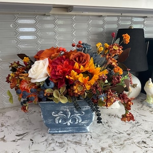May include: A floral arrangement featuring a mix of artificial flowers in shades of orange, red, yellow, and white, accented with dark blue berries and foliage, housed in a distressed blue rectangular planter with a raised scroll design.